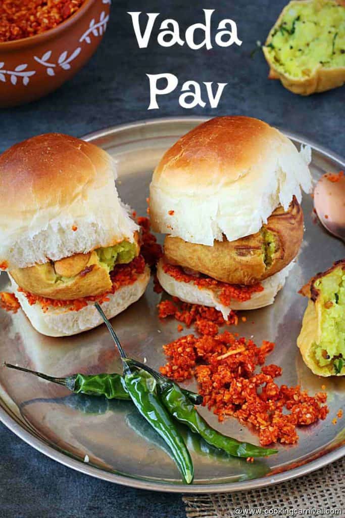2 vada pav in a steel plate with fried green chilies and dry garlic chutney on the side