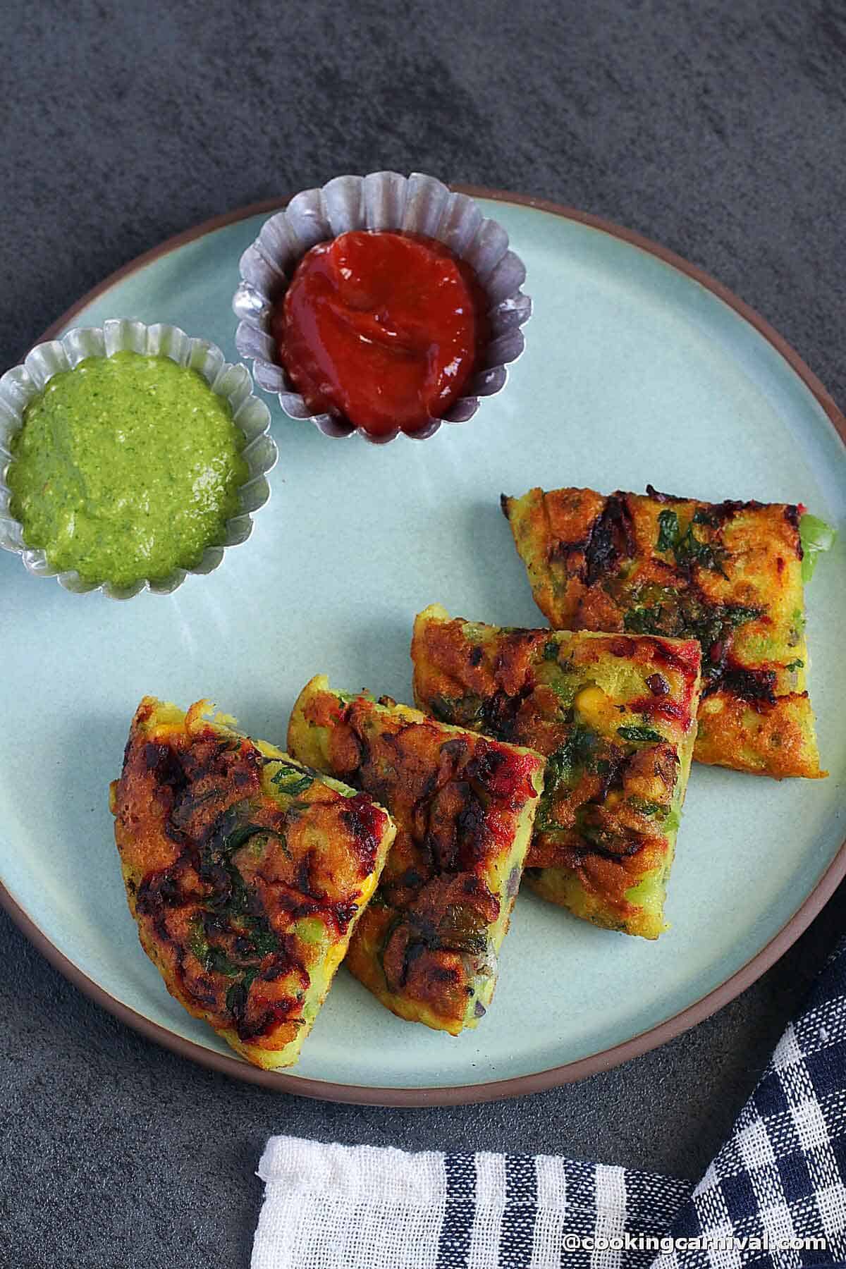 four pieces of moong dal pancake, green chutney and ketchup on a plate.